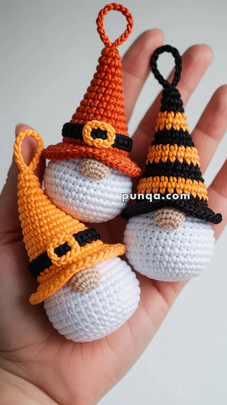 Three crocheted gnome ornaments in Halloween colors, each with a white body and pointed hat, held in a person's hand. The hats are orange, yellow with a black belt, and orange with black stripes, all with embellishments resembling belts.