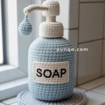 A crocheted soap dispenser bottle on a tiled bathroom countertop, featuring shades of cream and blue yarn, with the word 'SOAP' stitched on the front.