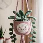 A crocheted hanging planter with a smiling face, featuring green crocheted leaves, hanging in a room with soft lighting.