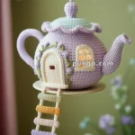 A decorative teapot-shaped hanging ornament made of crocheted material, featuring a miniature door and window, with a colorful ladder made of yarn leading up to it, set against a blurred green background.
