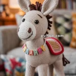 A crocheted donkey wearing a colorful saddle and a necklace with the letters spelling 'HAPPY', standing on a wooden stool in a cozy living room setting.
