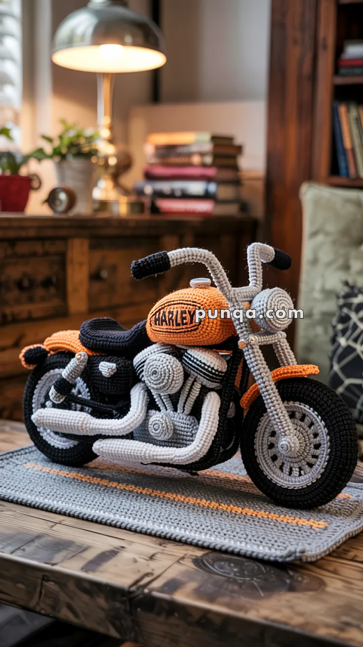 A detailed knitted motorcycle model in orange and black displayed on a wooden table, with books and a lamp in the background, creating a cozy interior setting.