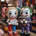 Harley and Joker crocheted dolls resembling iconic comic villains are standing on a wooden table. The doll on the left has blonde hair styled in pigtails, wearing a colorful outfit, and is holding a sign that reads 'Double Trouble.' The doll on the right has green hair, wearing a purple suit, and is holding a flower. The background shows a cozy room with light bulbs and yarn.