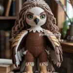 A crocheted plush Harpy toy resembling an anthropomorphic owl with large green eyes, brown feathers, and a white and brown body, sitting on a wooden stool with books in the background.