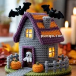 A crocheted Halloween-themed house with lit windows, featuring a purple roof and bats on top, a ghost at the door, pumpkins, and a gray picket fence, set on a wooden table with candles in the background.