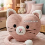 A heart-shaped, knitted pink cat pillow with white accents sits on a fuzzy pink blanket, featuring black eyes, a pink heart nose, and whiskers; the background shows a softly lit room with pastel decor and a teddy bear on a shelf.