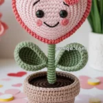 A crochet stuffed toy featuring a heart-shaped pink face with smiling eyes and cheeks, a flower stem, and two leaves, all set in a crochet pot, surrounded by paper heart decorations.