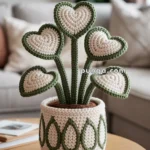 A crochet plant with heart-shaped leaves in beige and green, displayed in a matching decorative crochet pot on a wooden table in a living room setting.