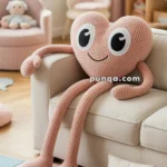 A plush heart-shaped cushion with smiling face and long arms and legs sits on a sofa in a cozy room with pastel decor, including a small pillow embroidered with "Dream Big" on the floor beside a colorful puzzle mat.