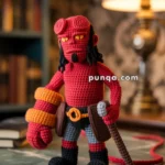 A crocheted doll of a red demon-like character with yellow eyes, black pants, and red and orange accessories, standing on a wooden table with books in the background.