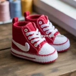 Red and white crocheted baby sneakers on a wooden surface, with colorful spools of thread in the background.