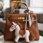 A brown leather handbag with a gold clasp and a decorative silk scarf tied around the handle, featuring a white and brown crocheted horse charm hanging from it, placed on a round marble table in a stylish room with a blurred background.