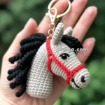 A crocheted horse head keychain with a red bridle, held in a hand against a blurred green background. The keychain has a golden clasp and the horse features a black mane.