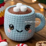 A cute crocheted mug with a smiling face, brown top resembling hot chocolate, and topped with crocheted marshmallows. It is surrounded by crocheted Christmas-themed decorations, including a candy cane and cookies, set on a wooden table.