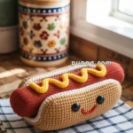 Crocheted plush resembling a hot dog with mustard, featuring a smiling face and black eyes, placed on a checkered cloth on a wooden countertop, with decorative kitchen containers in the background.