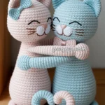 Two crochet cat dolls, one pink and one blue, are embracing each other while smiling, with their tails forming a heart shape on a wooden surface.