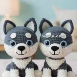 Two gray and white crocheted husky dog plush toys sitting side by side on a bed with a blurred background of a lamp and framed picture.