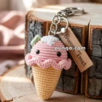 A handcrafted keychain featuring a crocheted ice cream cone with pink ice cream and black eyes, set on a wooden background. The keychain includes a metal ring and a small tag labeled "Sweet Treat."