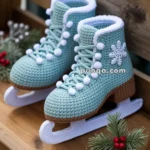 A pair of crocheted ice skates with light blue boots, white laces, and a snowflake motif, displayed on a wooden surface with a festive pine sprig and the words 'Happy Holidays.'