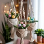 Three macramé plant hangers with crocheted plants and flowers in different colors hanging from a wooden rod in a cozy, well-lit room.