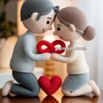 A crochet couple sits on a wooden table, kneeling face to face, holding a red infinity symbol between them.