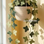 A crocheted hanging plant with green leaves in various shades, displayed in a beige basket near a sunlit window.