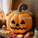 A crocheted pumpkin with a jack-o'-lantern face and a curling brown stem, surrounded by crocheted autumn leaves and candy corn decorations, placed on a wooden shelf next to a small lit candle.