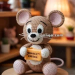 A crocheted mouse holding a block labeled "Double Cheese," sitting on a wooden stool in a cozy room.