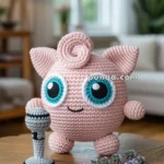 A crocheted pink toy character with large blue eyes and curly pink hair, holding a crocheted microphone, is placed on a wooden table with a lavender sprig in a cozy living room setting.
