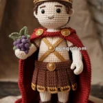 A crocheted doll dressed as a Roman soldier, wearing a brown skirt, red cape, and golden crown, holding a crocheted bunch of grapes. The doll stands on a cloth with the words "VENI VIDI VICI."