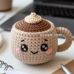 A crocheted plush mug with a smiling face and rosy cheeks, featuring a swirl pattern on top resembling foam, set on a desk beside a notebook, pens, and a plant.
