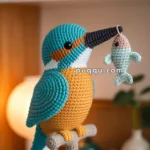 A crocheted kingfisher bird perched on a branch, holding a small crocheted fish in its beak, with a blurred indoor background.