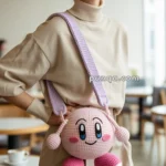 A person smiling while holding a pink crocheted bag shaped like a character with large eyes and feet, wearing a beige turtleneck sweater.