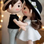 Crocheted dolls of a couple standing close together, with the male doll wearing a beige hat, black shirt, and pale green pants, and the female doll wearing a black hat with a white flower and a white dress, set against a softly lit background.