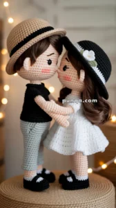 Crocheted dolls of a couple standing close together, with the male doll wearing a beige hat, black shirt, and pale green pants, and the female doll wearing a black hat with a white flower and a white dress, set against a softly lit background.