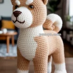 A crocheted dog resembling a Shiba Inu, made with tan and white yarn, standing on a wooden surface in a room with a couch in the background.