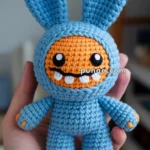 A handmade crochet doll with blue body and large ears, featuring an orange face with large eyes and two prominent teeth, held in a person's hand.