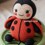 A crocheted ladybug amigurumi sitting on a green knitted leaf, featuring a red body with black spots and a smiling face.