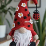 Amigurumi gnome with a red hat adorned with black dots and white flowers, featuring a long white beard and a small crocheted ladybug hanging from the hat, held in a person's hand.