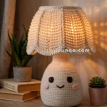 A crochet lamp with a smiling face design, featuring a textured lampshade, sits on a wooden table beside potted plants and books.