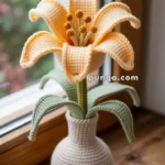 A crocheted orange lily with green leaves in a matching crocheted vase, placed on a wooden windowsill.