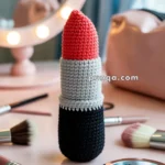 A crocheted red lipstick with a black base and gray middle section, placed among makeup brushes and a compact mirror on a vanity table.