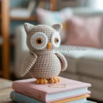 A crocheted owl toy with large round eyes and a beige body sits atop two stacked, pastel-colored books on a wooden table in a cozy living room setting.