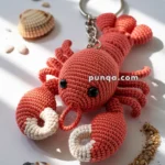 A crocheted lobster keychain with large black eyes and detailed claws, surrounded by small seashells and sand on a white surface.