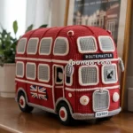 A crocheted model of a red double-decker bus with the word "ROUTEMASTER" on the front, featuring detailed windows and wheels, sitting on a wooden surface with a framed London poster in the background.