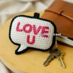 A crocheted speech bubble keychain with the words "LOVE U" in pink letters, placed on a wooden surface next to a notebook and two keys.