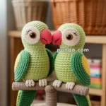 Two crocheted parrot figures with green bodies, yellow wing patches, and red beaks, perched on a yarn-covered stand, with a basket of colorful yarn balls in the blurred background.