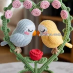 A crocheted artwork featuring two birds, one blue and one yellow, perched on a green heart-shaped frame adorned with pink roses. The birds are kissing, positioned above a red crocheted rose, all set on a brown circular base.