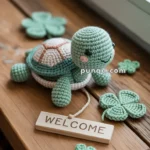A crocheted turtle toy sits on a wooden surface surrounded by crocheted four-leaf clovers and a small "Welcome" sign.
