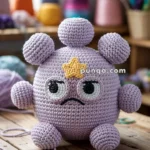 A crocheted plush resembling a purple cartoon-style character with a star on its forehead, large eyes, and several round appendages, sitting on a wooden table with various yarns and crafting supplies in the background.